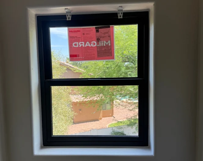 A newly installed black framed window with a red Milgard sticker and two white mounting brackets at the top.
